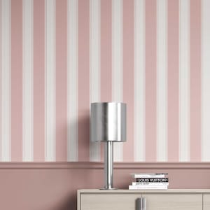 May include: A silver table lamp with a cylindrical shade sits on a light wood cabinet. The background features a pink and white striped wallpaper. Books with the titles "LOUIS VUITTON" and "pierre cardin" are stacked on the cabinet.