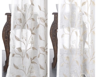 May include: Two white sheer curtains with a delicate embroidered floral pattern in a light brown color. The curtains are hanging from a rod and are partially drawn, revealing a glimpse of a wooden table behind them.