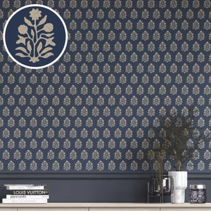 Op de afbeelding: Marineblauwe behang met een herhalend bloemmotief in een lichtbeige kleur. Een close-up toont het bloemmotief in een cirkel. Het behang is geïnstalleerd op een muur met een donkerblauwe lijst en een witte plank.