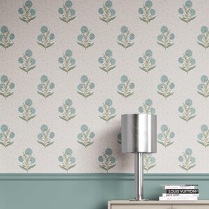 Op de afbeelding: Een behangpatroon met een herhalend bloemenontwerp in blauw en groen tegen een crèmekleurige achtergrond. De afbeelding toont ook een zilveren lamp, een turquoise muurbekleding en een stapel boeken.
