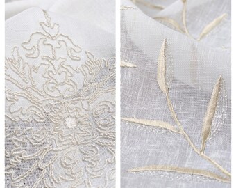 May include: Close-up of two white sheer curtains with embroidered designs. One curtain features a large, intricate floral pattern, while the other has a simple, repeating leaf design. Both designs are in a light beige color.