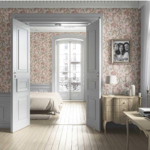 May include: A bedroom interior with light wood flooring and floral wallpaper in pink and green tones. The room features a bed, a wooden dresser, a framed family photo, and a table with a lamp and candles. White doors and a window with a balcony are also visible.