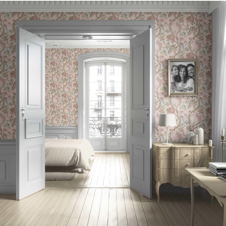 May include: A bedroom interior with light wood flooring and floral wallpaper in pink and green tones. The room features a bed, a wooden dresser, a framed family photo, and a table with a lamp and candles. White doors and a window with a balcony are also visible.