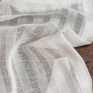 Pode incluir: Close-up de um tecido branco semitransparente com textura tecida. O tecido está drapeado, criando dobras e sombras suaves. A palavra "ORELIA" é visível no topo. O tecido parece ser linho ou material semelhante.