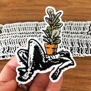 Puede incluir: Parche bordado en blanco y negro de una persona acostada con una planta en maceta sobre el estómago. La planta tiene hojas verdes y una maceta naranja.