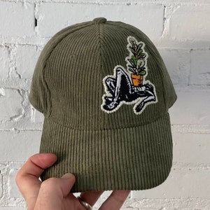 May include: A green corduroy baseball cap featuring a black and white embroidered patch of a person sitting with a potted plant on their head.