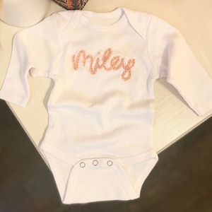 Op de afbeelding: Een wit babyrompertje met lange mouwen en de naam Miley geborduurd in licht perzikkleurig garen op de borst. Het kledingstuk heeft een ronde halslijn en drie metalen drukknopen aan de onderkant voor gemakkelijke toegang.