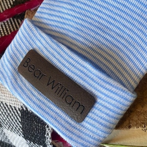 Op de afbeelding: Een baby beanie met lichtblauwe en witte strepen, met een bruin leren label met de naam "Bear William". De opgevouwen beanie ligt op een kurken ondergrond. Een zwart-wit geblokt patroon is zichtbaar op de achtergrond.