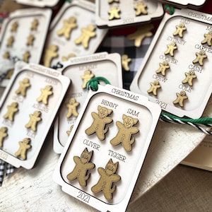 Op de afbeelding: Een set houten kerstversieringen met gingerbread-poppetjes en dierlijke ontwerpen. Elke versiering heeft een andere naam en het jaar 2021. De versieringen zijn versierd met gouden verf en hebben een witte achtergrond.