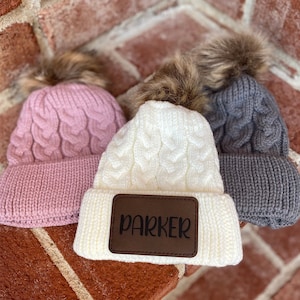 Op de afbeelding: Drie gebreide mutsen met nep bont pompoenen. E&eacute;n muts is roze, &eacute;&eacute;n is wit met een bruin leren patch waarop "PARKER" staat, en &eacute;&eacute;n is grijs.