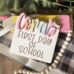 May include: A white wooden sign with the text "Coras First Day of School" in brown lettering. The name "Coras" is in rainbow colors and is layered on top of the sign.