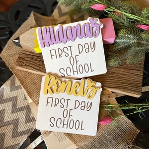 May include: Two white signs with the words "First Day of School" in brown lettering. The top sign has the name "Hildana's" in purple lettering. The bottom sign has the name "Ronel's" in gold lettering. The signs are on a wooden surface with a burlap ribbon and a pencil.