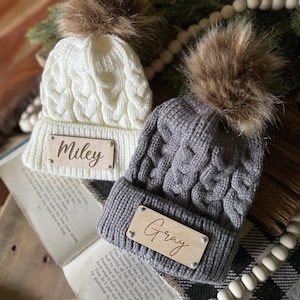 Op de afbeelding: Twee gebreide wintermutsen met nep bont pompoenen. De mutsen zijn grijs en wit met houten naamschildjes met de namen "Miley" en "Gray".