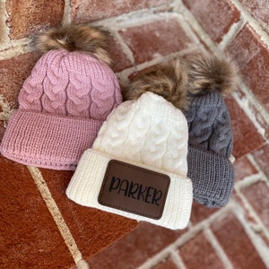 Op de afbeelding: Drie gebreide wintermutsen met nep bont pompoenen. De witte muts heeft een bruin leren patch met de naam "Parker" erop.