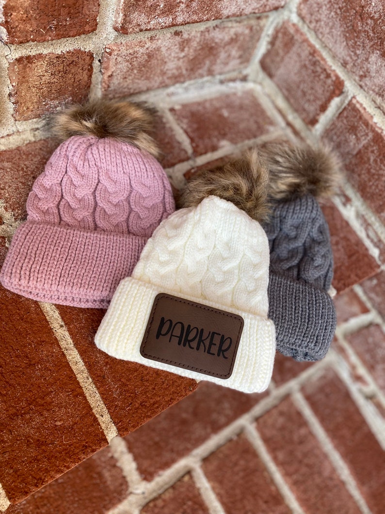 Op de afbeelding: Drie gebreide wintermutsen met nep bont pompoenen. De witte muts heeft een bruin leren patch met de naam "Parker" erop.