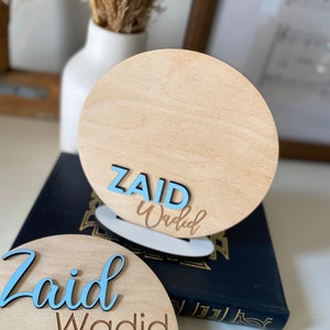 May include: Two wooden round signs with the name "Zaid Wadid" written in blue and brown lettering. The signs are on a dark blue surface.