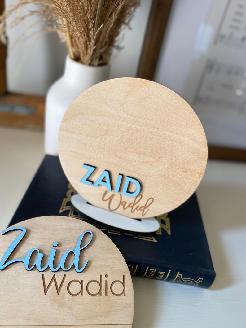 May include: Two wooden round signs with the name "Zaid Wadid" written in blue and brown lettering. The signs are on a dark blue surface.
