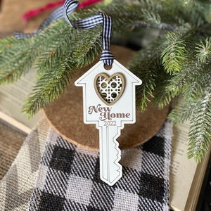 May include: White wooden key-shaped ornament with a house cutout and a brown heart in the center. The ornament says "New Home 2022".