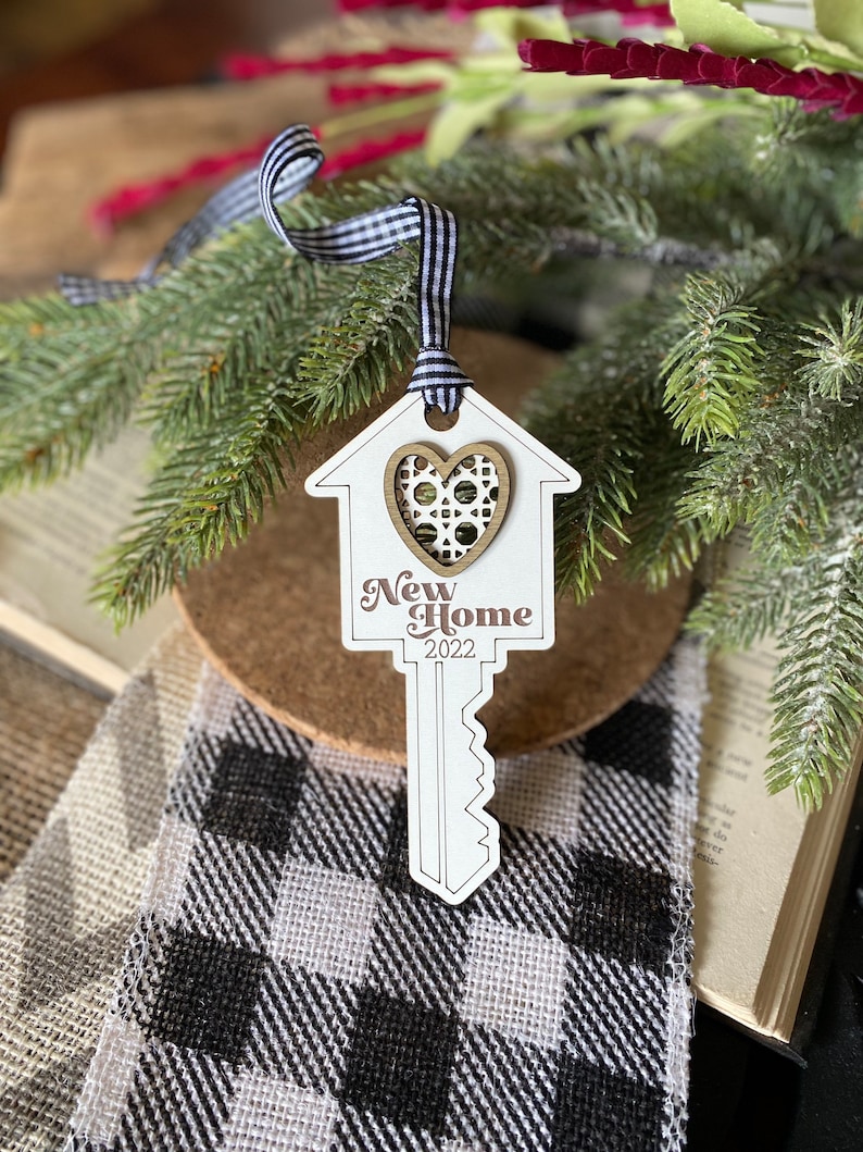 May include: White wooden key-shaped ornament with a house cutout and a brown heart in the center. The ornament says "New Home 2022".