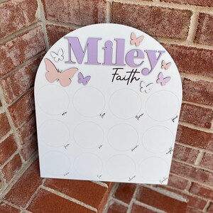 May include: White wooden arch with the names "Miley Faith" in purple lettering. The arch has 12 circles with numbers one through twelve written inside. There are pink and purple butterflies on the arch.
