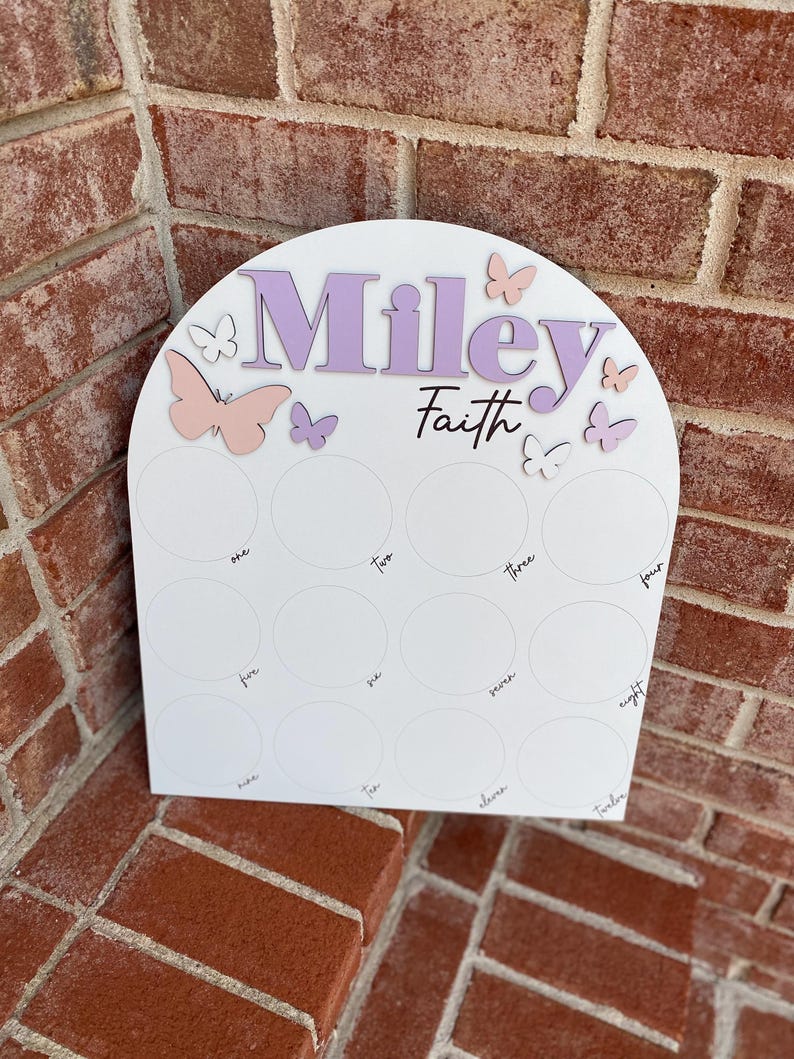 May include: White wooden arch with the names "Miley Faith" in purple lettering. The arch has 12 circles with numbers one through twelve written inside. There are pink and purple butterflies on the arch.