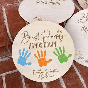 May include: A round wooden sign with the text "Best Daddy Hands Down!" in a cursive font. Three colorful handprints are displayed below the text. The names "Natalie Sebastian & Sullivan" are written below the handprints.