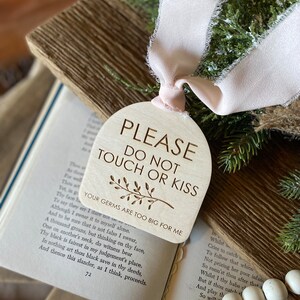 May include: A light-colored wooden arch-shaped sign with the engraved text PLEASE DO NOT TOUCH OR KISS, a small branch illustration, and the text YOUR GERMS ARE TOO BIG FOR ME, finished with a pale pink frayed ribbon tied at the top.