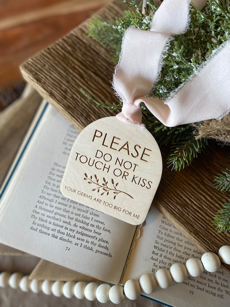May include: A light-colored wooden arch-shaped sign with the engraved text PLEASE DO NOT TOUCH OR KISS, a small branch illustration, and the text YOUR GERMS ARE TOO BIG FOR ME, finished with a pale pink frayed ribbon tied at the top.