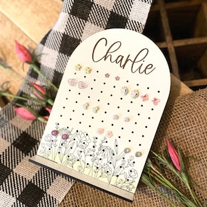 May include: White wooden earring holder with black polka dots and a floral design. The holder has a rounded top with the name "Charlie" written in cursive. The holder is filled with colorful earrings.