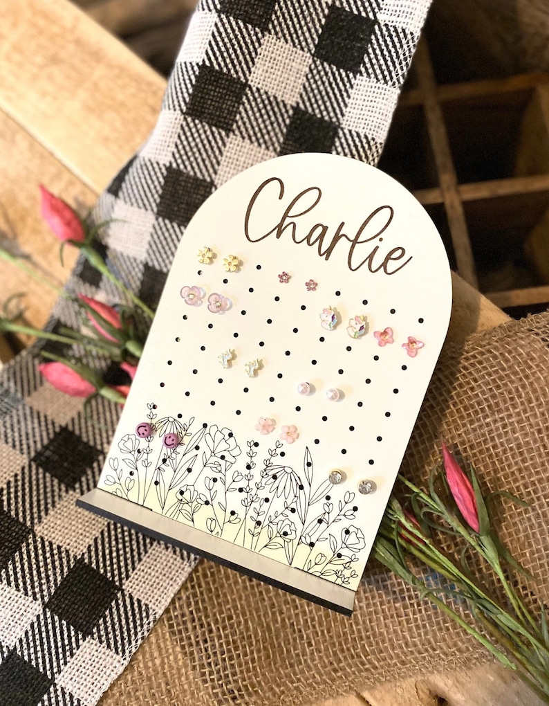 May include: White wooden earring holder with black polka dots and a floral design. The holder has a rounded top with the name "Charlie" written in cursive. The holder is filled with colorful earrings.