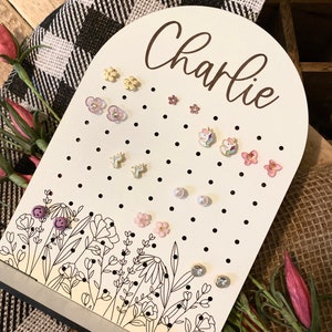 May include: White wooden earring holder with the name "Charlie" written in brown lettering. The holder has a floral design and is decorated with small black dots. The holder is filled with various colorful earrings, including flowers, stars, and unicorns.
