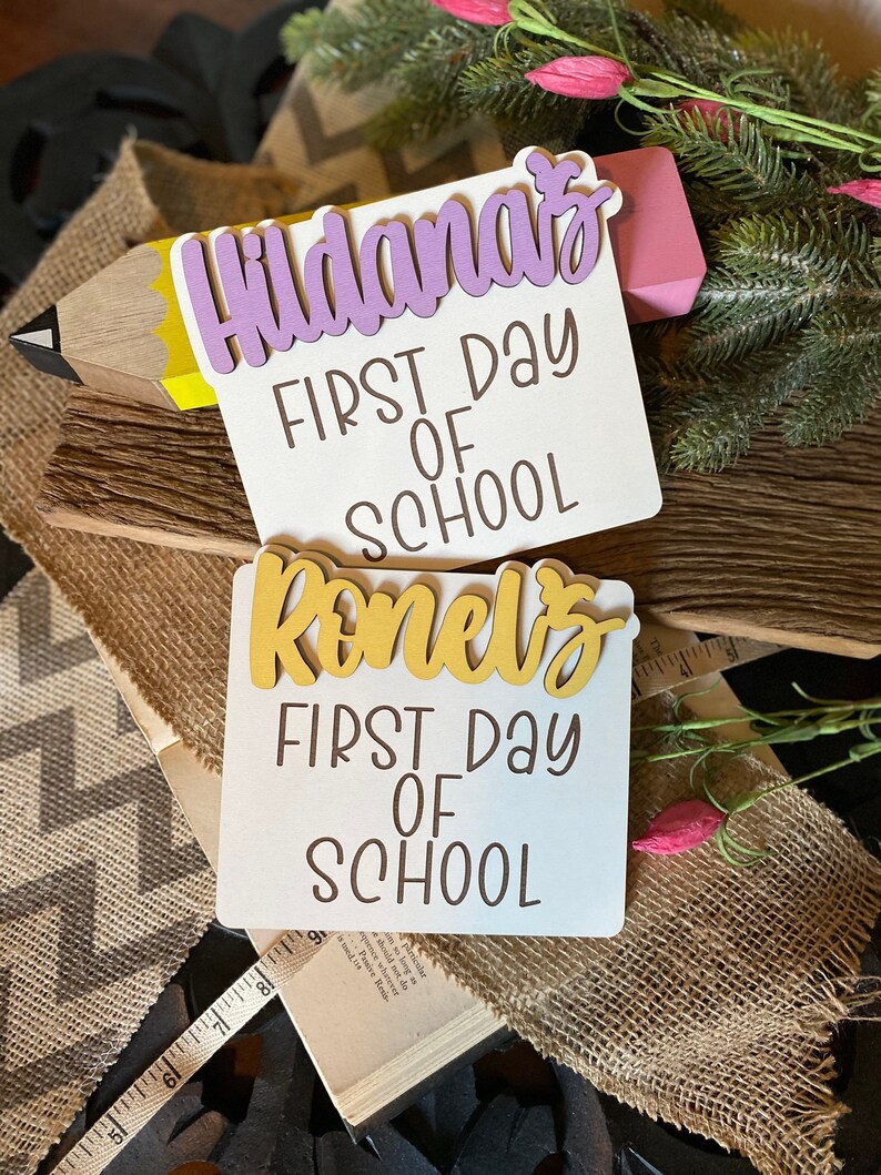 May include: Two white signs with the text "First Day of School" in brown lettering. The top sign has a purple name cutout that says "Hildana's". The bottom sign has a yellow name cutout that says "Reneva's".