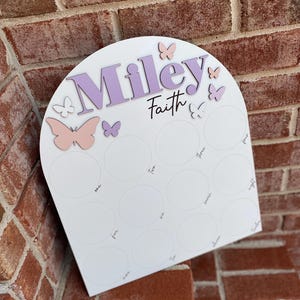 May include: White arch-shaped board with the names "Miley" and "Faith" written in purple. The board has 12 circles with numbers one through twelve written inside. There are pink and purple butterflies around the names.