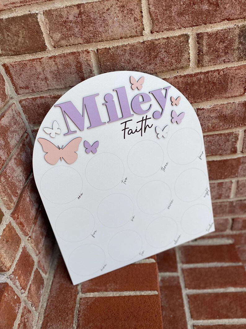 May include: White arch-shaped board with the names "Miley" and "Faith" written in purple. The board has 12 circles with numbers one through twelve written inside. There are pink and purple butterflies around the names.