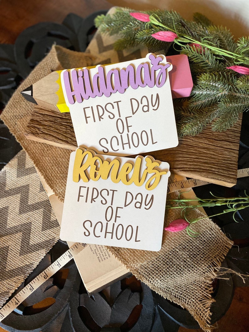 May include: Two white signs with the words "First Day of School" in brown lettering. The top sign has the name "Hildana's" in purple lettering. The bottom sign has the name "Ronel's" in gold lettering. The signs are on a wooden surface with a burlap ribbon and a pencil.