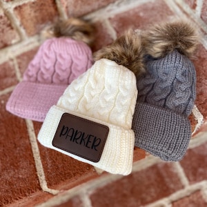 Op de afbeelding: Drie gebreide mutsen met nep bont pompoenen. E&eacute;n muts is roze, &eacute;&eacute;n is wit met een bruin leren patch waarop "Parker" staat, en &eacute;&eacute;n is grijs.