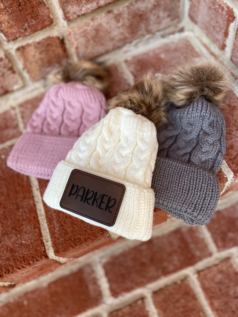 Op de afbeelding: Drie gebreide mutsen met nep bont pompoenen. E&eacute;n muts is roze, &eacute;&eacute;n is wit met een bruin leren patch waarop "Parker" staat, en &eacute;&eacute;n is grijs.