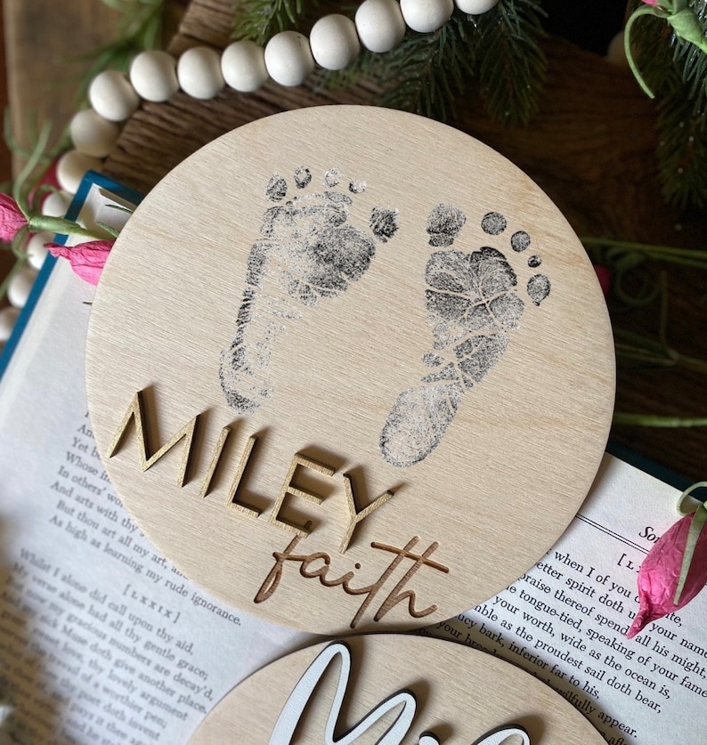 May include: A wooden round ornament with two baby footprints in black ink. The names "MILEY" and "faith" are written in gold lettering.