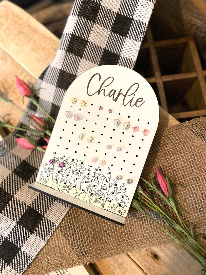 May include: A white wooden earring holder with a floral design and black polka dots. The holder has a cutout arch at the top with the name "Charlie" written in cursive. The holder is filled with various colorful earrings.