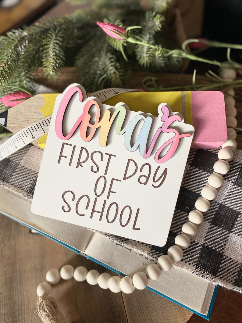 May include: A white wooden sign with the text "Coras First Day of School" in brown lettering. The name "Coras" is in rainbow colors and is layered on top of the sign.