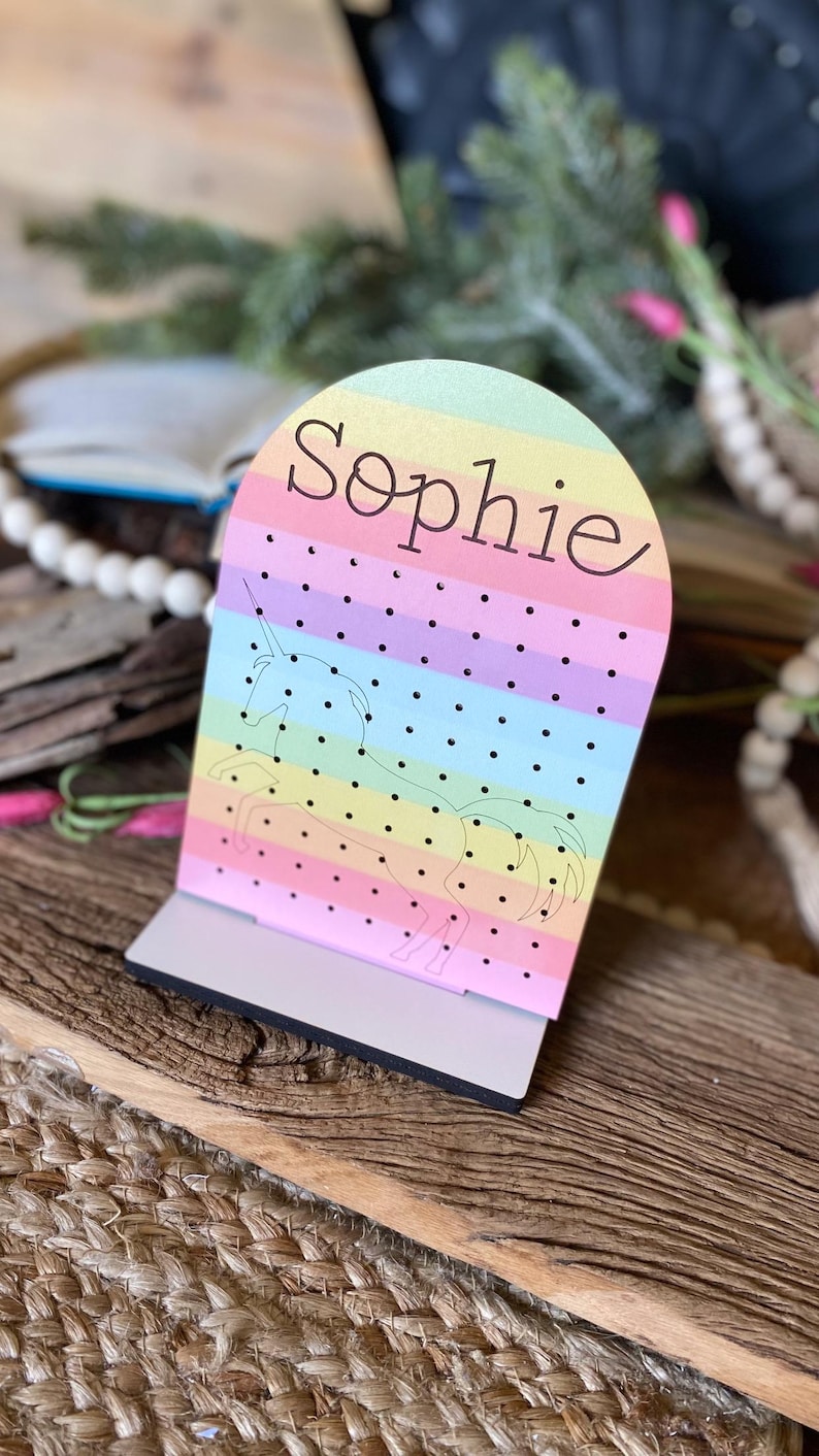 May include: A rainbow-colored wooden earring holder with a unicorn silhouette and the name "Sophie" printed on it.