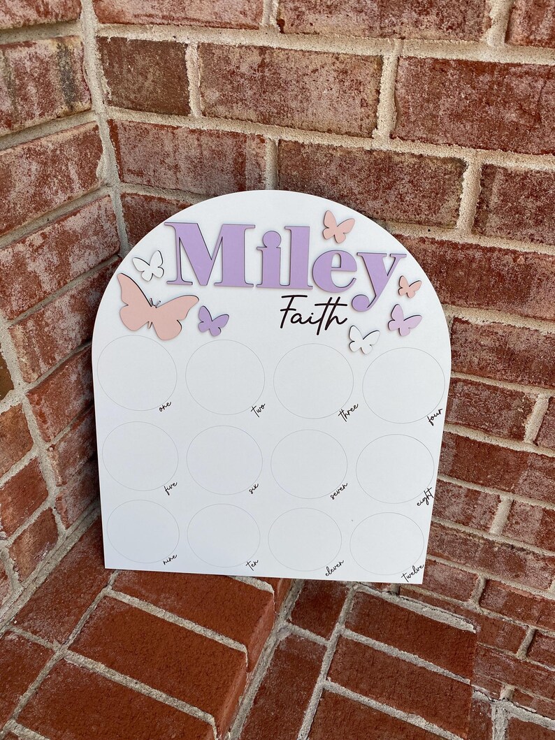 May include: A white arch-shaped wooden board with the names "Miley Faith" written in purple. The board has 12 circles with numbers one through twelve written in black. There are pink and purple butterflies around the names.