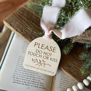 May include: A light-colored wooden arch-shaped sign with a pale pink frayed ribbon. The text reads: PLEASE DO NOT TOUCH OR KISS, YOUR GERMS ARE TOO BIG FOR ME, with a small engraved branch illustration centered below the main text.