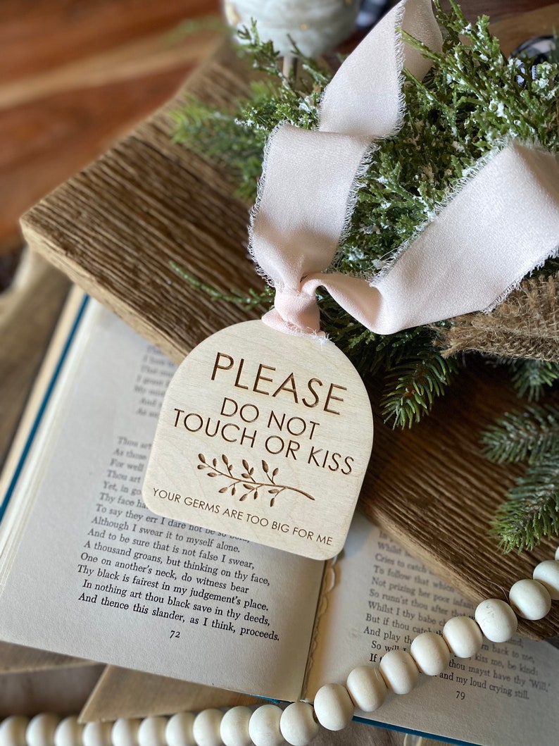 May include: A light-colored wooden arch-shaped sign with a pale pink frayed ribbon. The text reads: PLEASE DO NOT TOUCH OR KISS, YOUR GERMS ARE TOO BIG FOR ME, with a small engraved branch illustration centered below the main text.