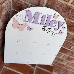 May include: White wooden arch with the name "Miley Faith" in purple lettering. The arch has 12 circles with numbers one through twelve written inside. There are pink and white butterflies on the arch.