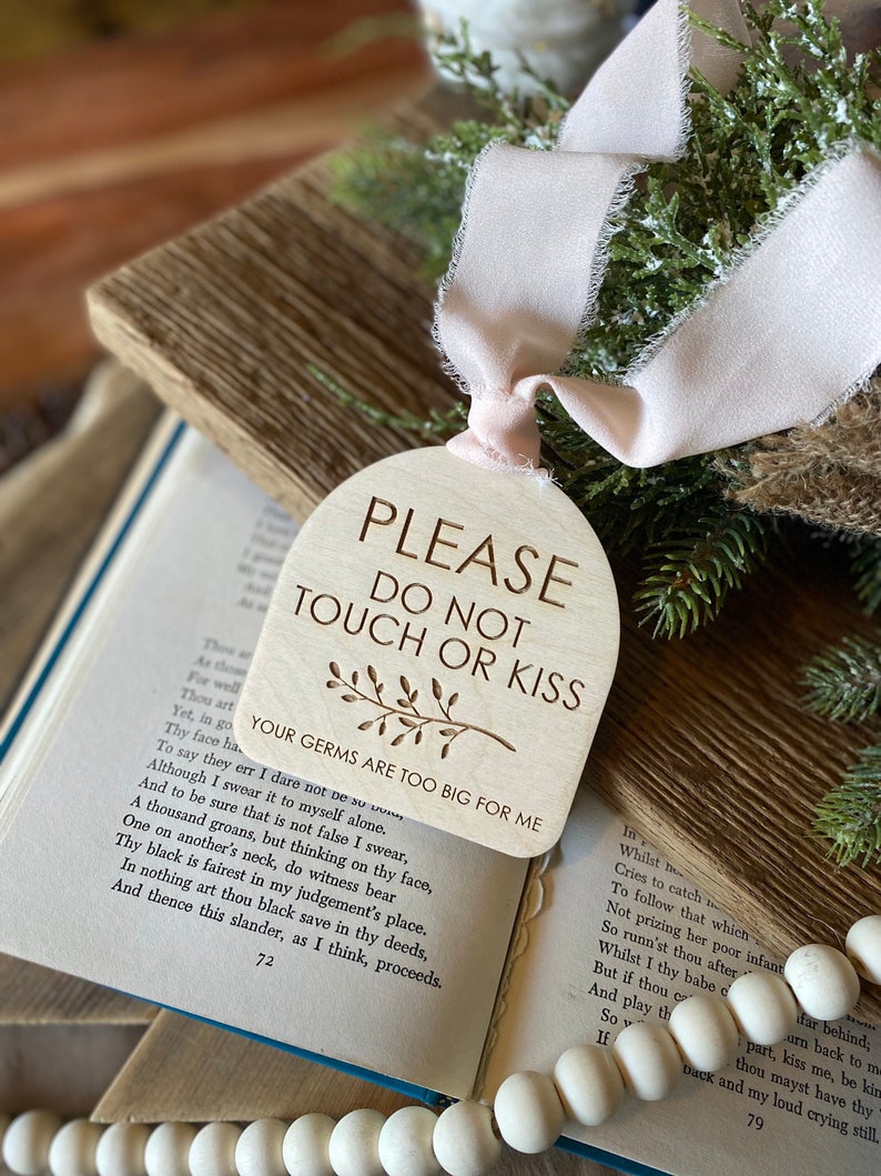 May include: An arched wooden sign with a light wood grain finish, featuring the engraved text "PLEASE DO NOT TOUCH OR KISS" and "YOUR GERMS ARE TOO BIG FOR ME" with a small leaf graphic. A pale pink frayed ribbon is tied through a hole at the top.