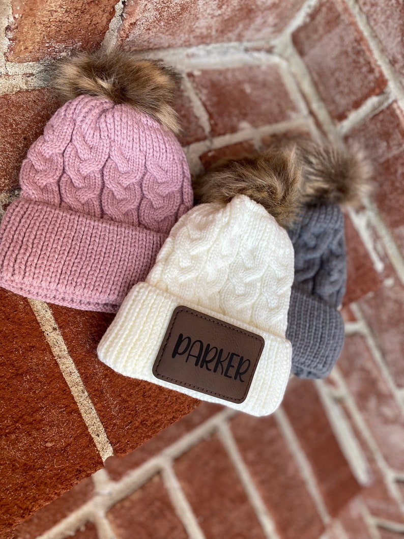 Op de afbeelding: Drie gebreide wintermutsen met nep bont pompoenen. E&eacute;n muts is roze, &eacute;&eacute;n is wit met een bruin leren patch waarop "Parker" staat, en &eacute;&eacute;n is grijs.