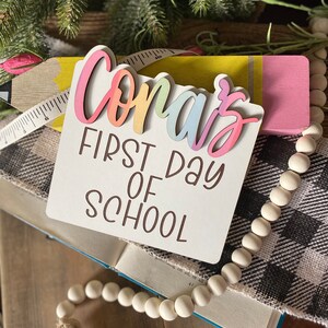 May include: A white wooden sign with the text "Conar's First Day of School" in brown lettering. The name "Conar's" is in rainbow colors and is layered on top of the sign.