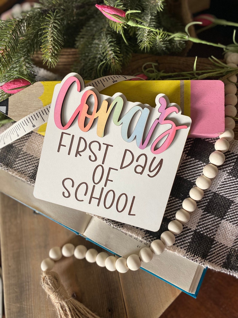 May include: A white wooden sign with the text "Conar's First Day of School" in brown lettering. The name "Conar's" is in rainbow colors and is layered on top of the sign.
