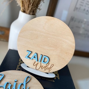 May include: Two wooden round signs with the name "Zaid Waqdid" written in blue and brown lettering. The signs are on a black surface.